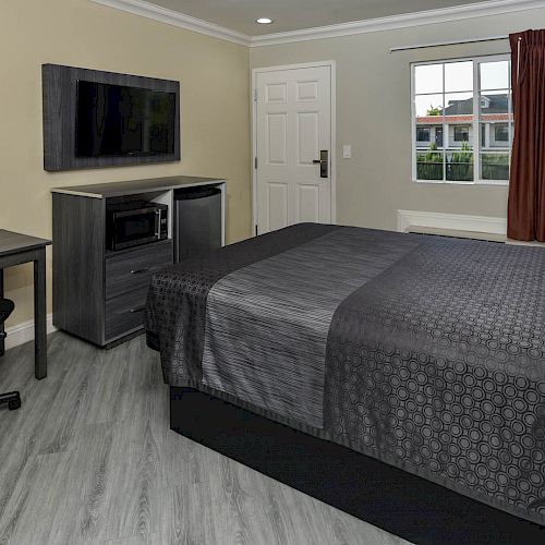 A modern hotel room with a bed, desk and chair, flat-screen TV, microwave, and a window with red curtains. Tan walls, wood flooring, and white door.