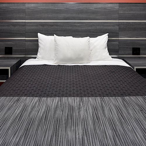 A neatly made bed with a black and gray headboard against a reddish wall, flanked by floating nightstands, and adorned with white pillows.