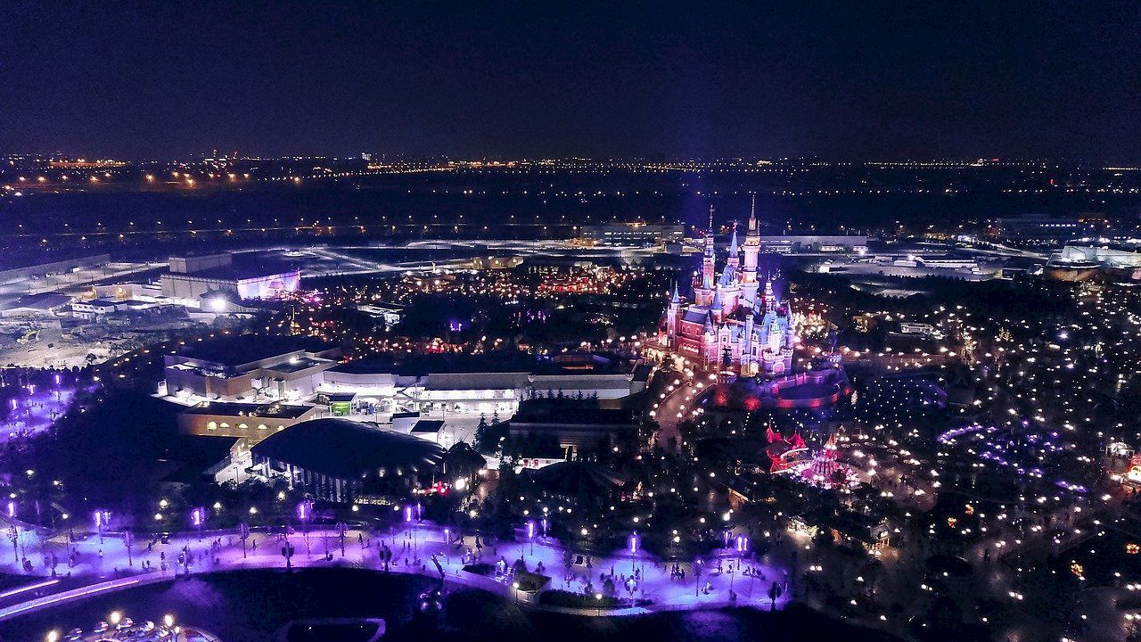 A night-time aerial view of a brightly lit theme park featuring a large, illuminated castle and various attractions, surrounded by city lights.