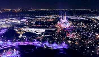 An aerial view of a theme park at night, beautifully illuminated with various lights and attractions, including a prominent castle.