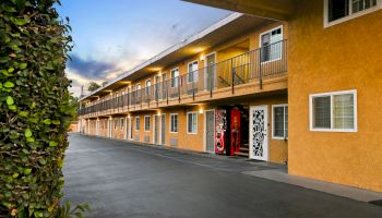 A two-story motel with outdoor corridors, a vending machine, orange exterior walls, windows, and doors, with a hedge on the left under a blue sky.