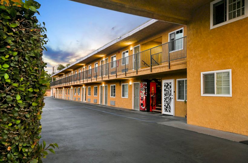 A two-story motel with outdoor corridors, a vending machine, orange exterior walls, windows, and doors, with a hedge on the left under a blue sky.