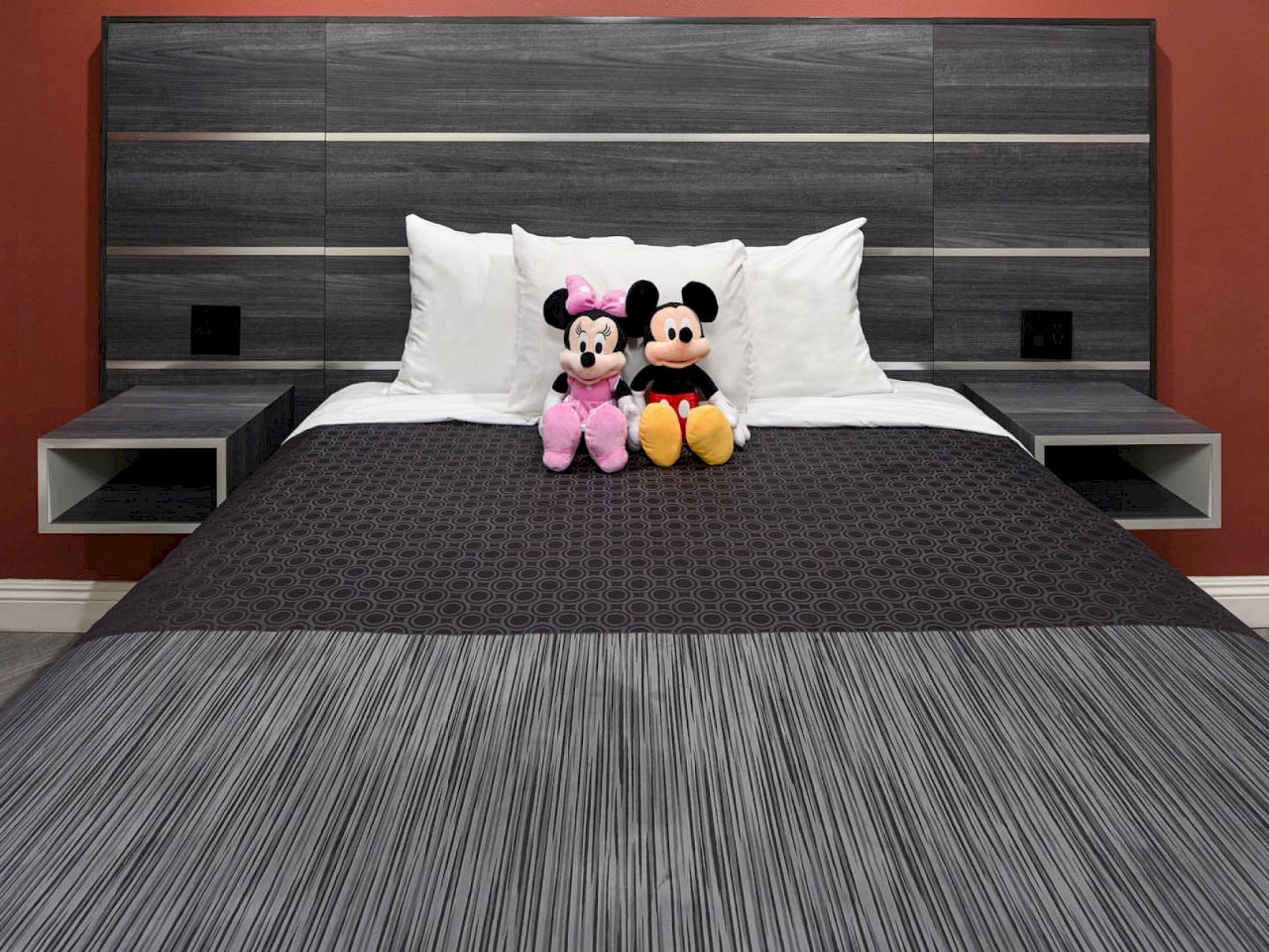 A bed with a dark wood headboard, white pillows, and two stuffed Mickey Mouse and Minnie Mouse dolls on top of the bedspread.
