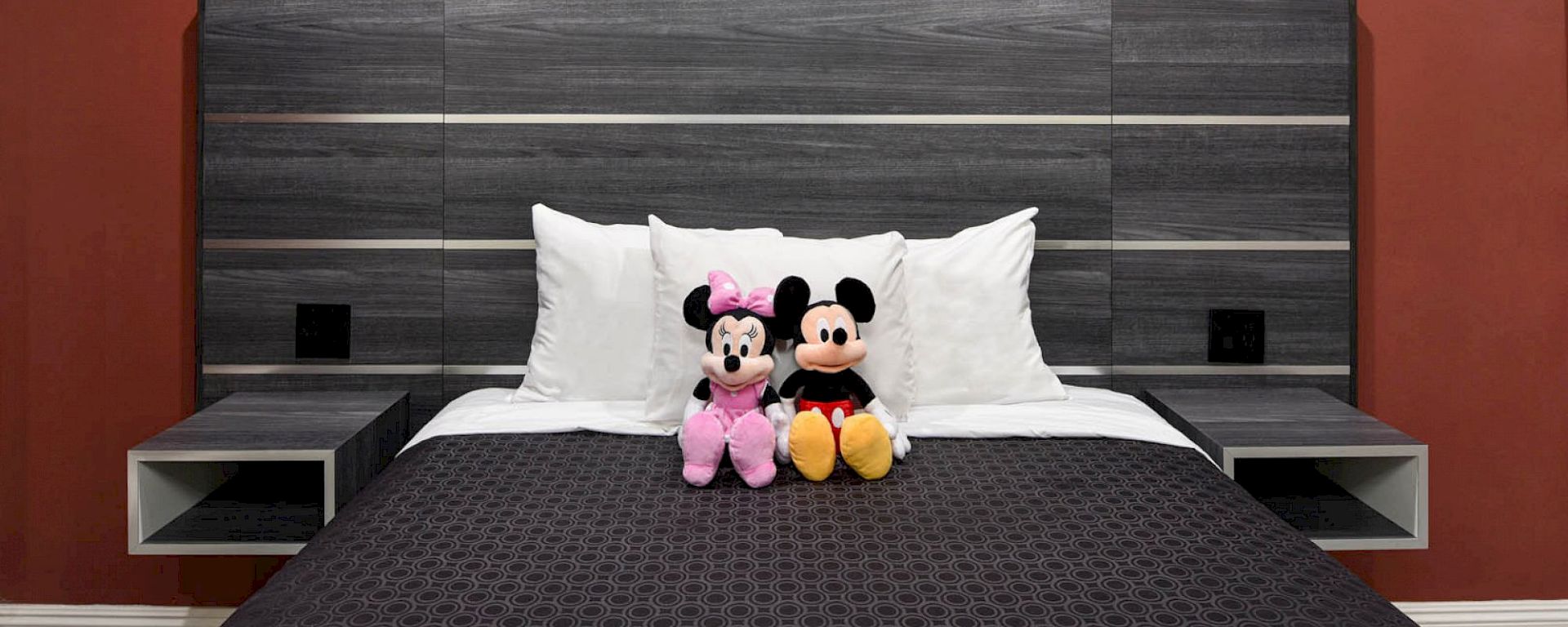 A bed with a dark wooden headboard, white pillows, and two stuffed Disney characters, Mickey and Minnie Mouse, sitting on it.