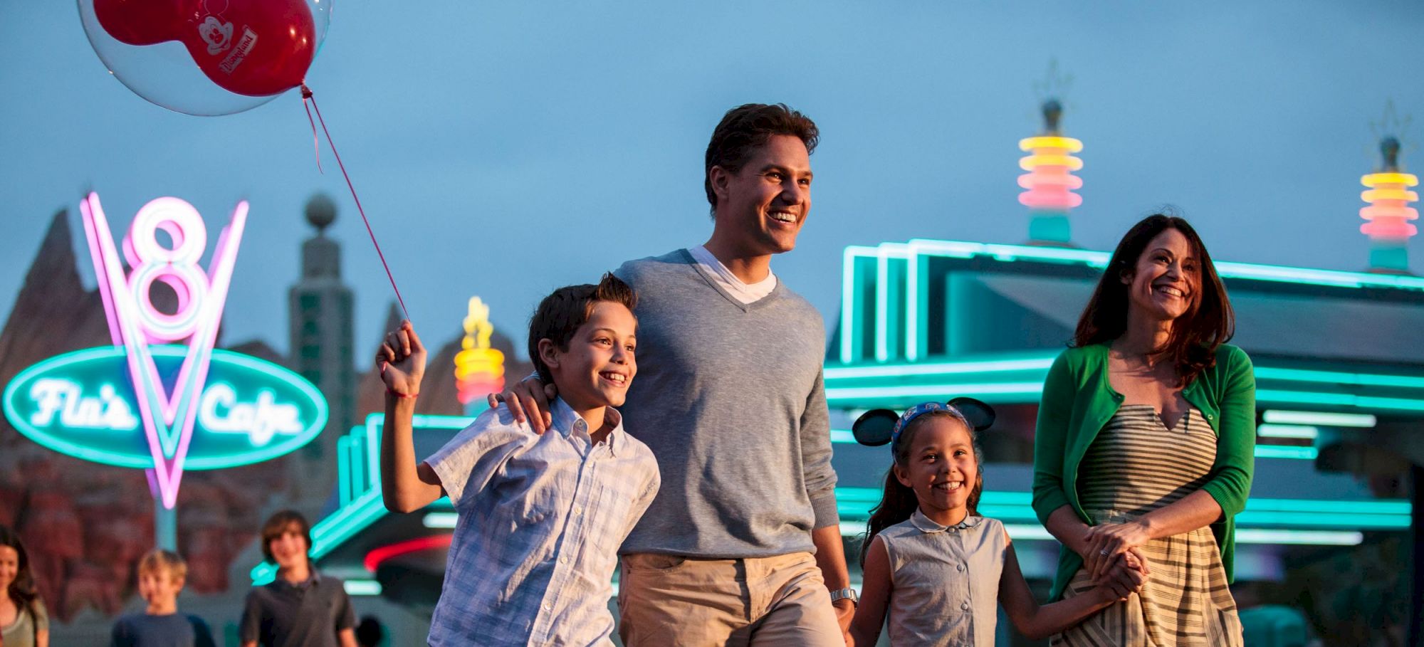 A family of four smiles while walking past a neon-lit cafe. One child holds a red balloon.