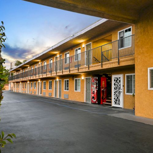 The image shows a two-story motel with exterior corridors and a vending machine. The building is painted yellow and has a hedge nearby.