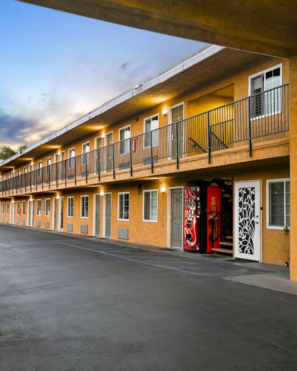 The image shows a two-story motel with exterior corridors and a vending machine. The building is painted yellow and has a hedge nearby.