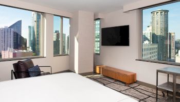 A modern hotel room with large windows offering a city view, a bed, lounge chair, wall-mounted TV, and minimalist furnishings.