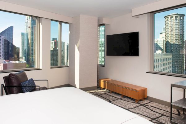 A wide-angle view of a hotel room with a bed, TV, chair, and table in front of large windows showing a city view.
