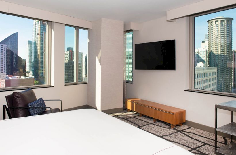 A modern hotel room with large windows offering a city view, a bed, lounge chair, wall-mounted TV, and minimalist furnishings.