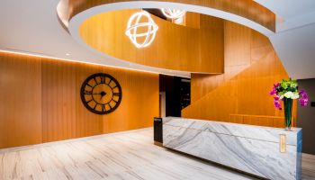 A modern interior with a large clock on a wooden wall, a curved wooden structure, marble reception desk, and decorative light fixtures.