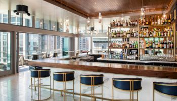 A modern bar with a wooden counter, high chairs, and a large selection of bottles on shelves. Large windows offer a view of the city skyline.