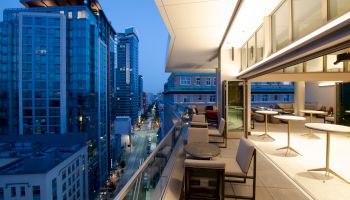Image of a modern city from a high-rise balcony, showing skyscrapers, a street below, and a sleek outdoor seating area with tables and chairs.
