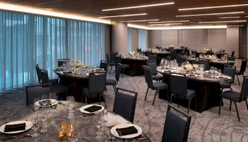 The image shows an elegantly set banquet hall with round tables, each adorned with glasses, plates, and black napkins, under modern lighting.