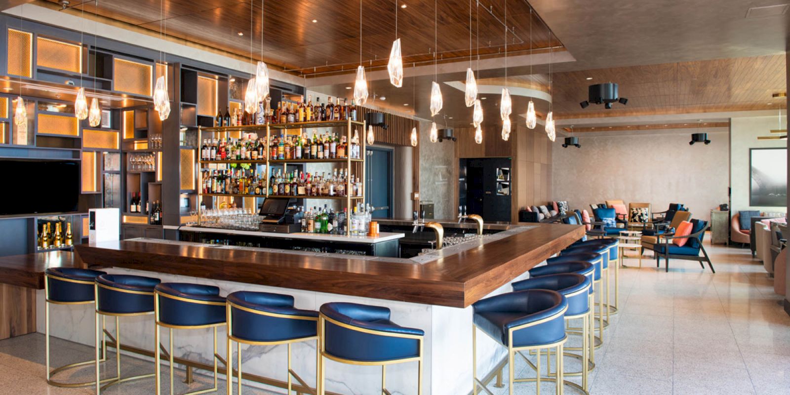The image shows a modern bar with a wooden countertop, blue chairs, a ceiling with hanging lights, and a well-stocked back shelf with various bottles.
