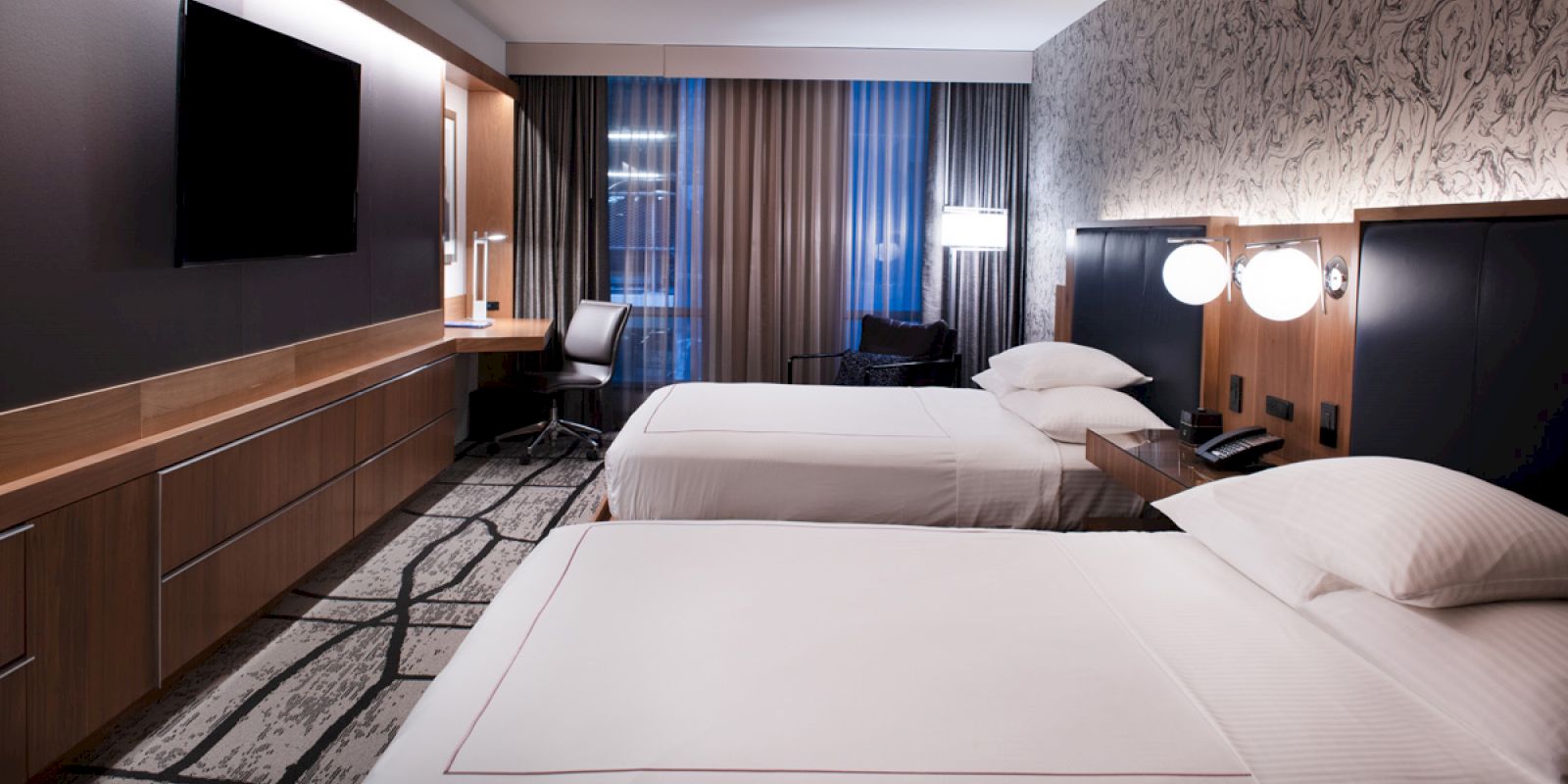 A modern hotel room with two neatly made beds, a wall-mounted TV, a desk with a chair, and large windows with curtains, well-lit and stylishly decorated.