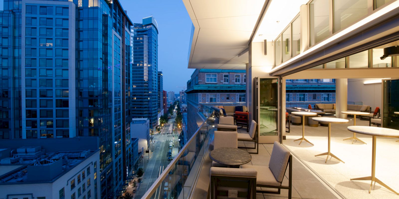 The image shows a modern cityscape from a high balcony with seating and tables, overlooking streets and tall glass buildings at dusk.