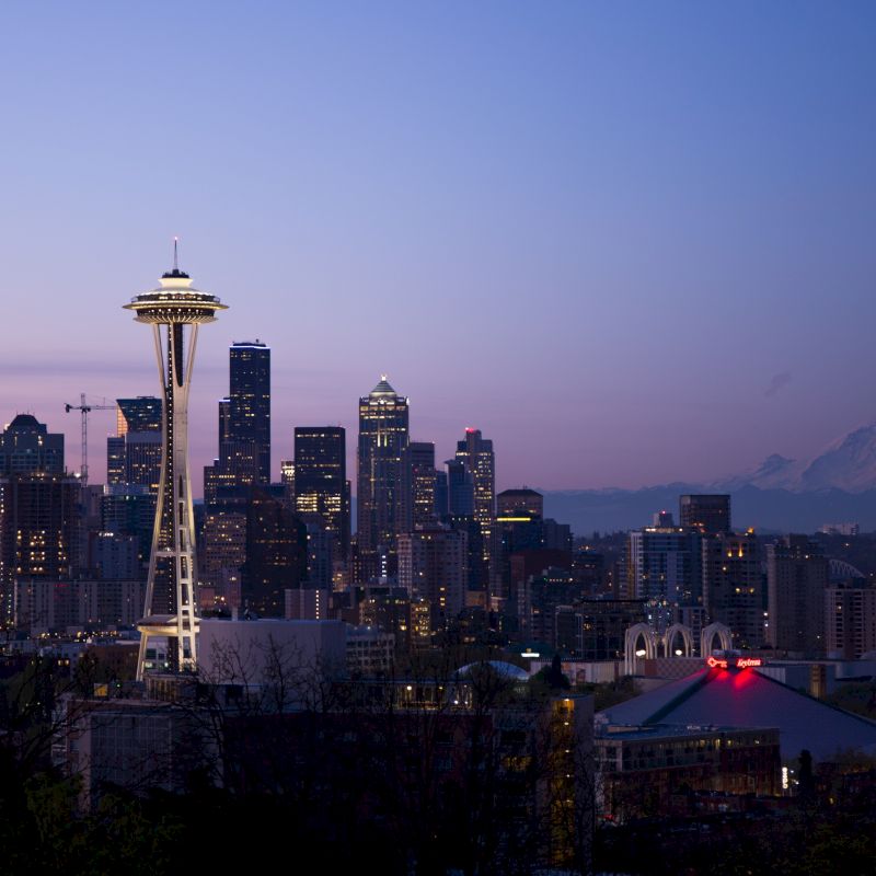 A twilight view of a city's skyline featuring a prominent tower, with a distant mountain and buildings illuminated against the fading sunlight.