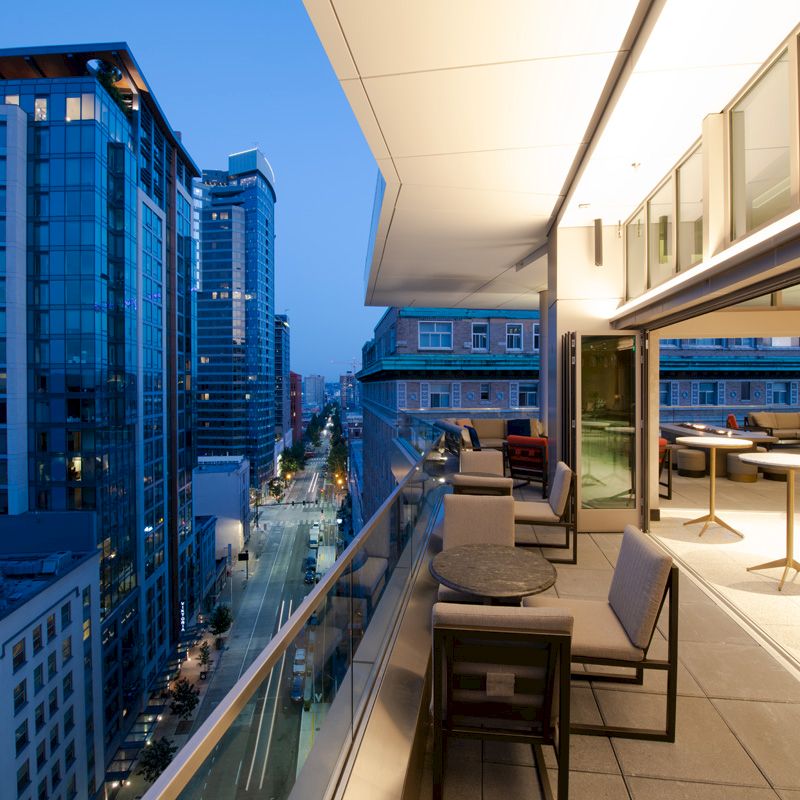 An urban balcony scene at dusk with modern high-rise buildings and streetlights visible, featuring outdoor seating and tables on a spacious deck.