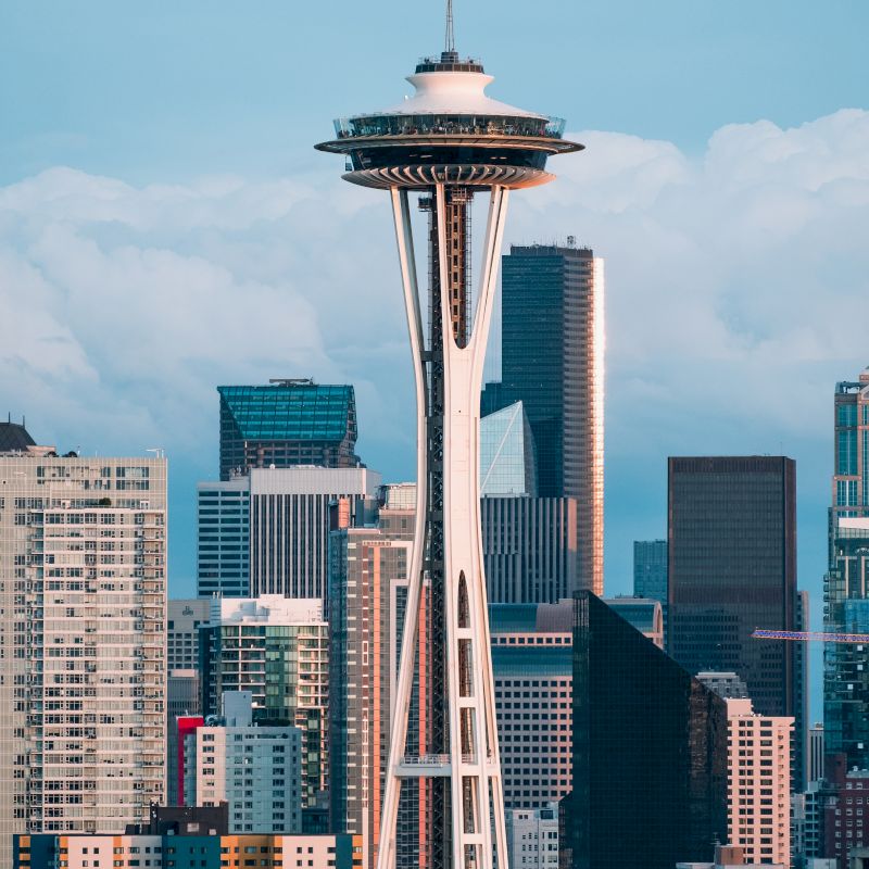The image shows the Space Needle with various skyscrapers in the background, situated in Seattle, Washington, under clear blue skies and light clouds.