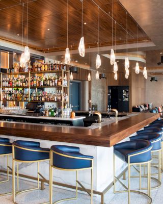 A modern bar features stylish blue barstools, a variety of liquor bottles on shelves, hanging lights, and a seating area with chairs and tables.