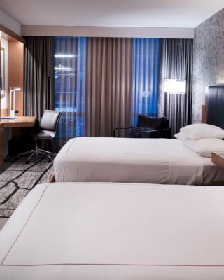 A modern hotel room features two neatly made beds, a wall-mounted TV, a desk with a chair, and large windows with curtains.