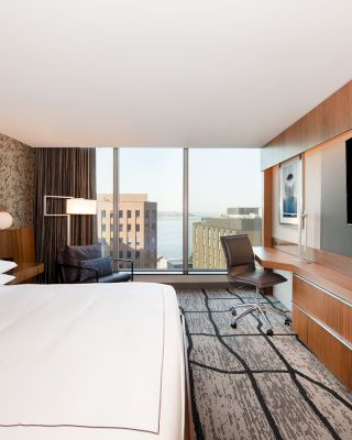 A modern hotel room features a large bed, wall-mounted TV, desk, chair, and a window with a city view.