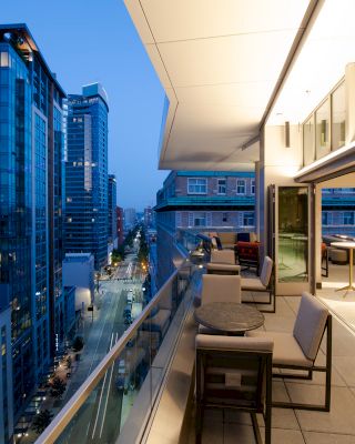 The image shows a modern cityscape with tall buildings at dusk, viewed from a stylish balcony with seating and tables.