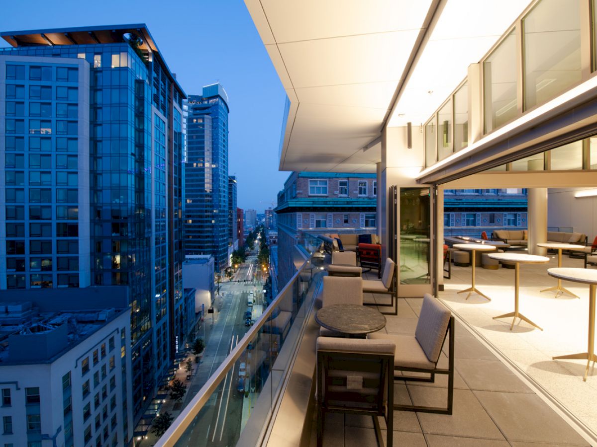A modern rooftop terrace with seating overlooks a cityscape with tall buildings at dusk. The terrace features tables, chairs, and ambient lighting.