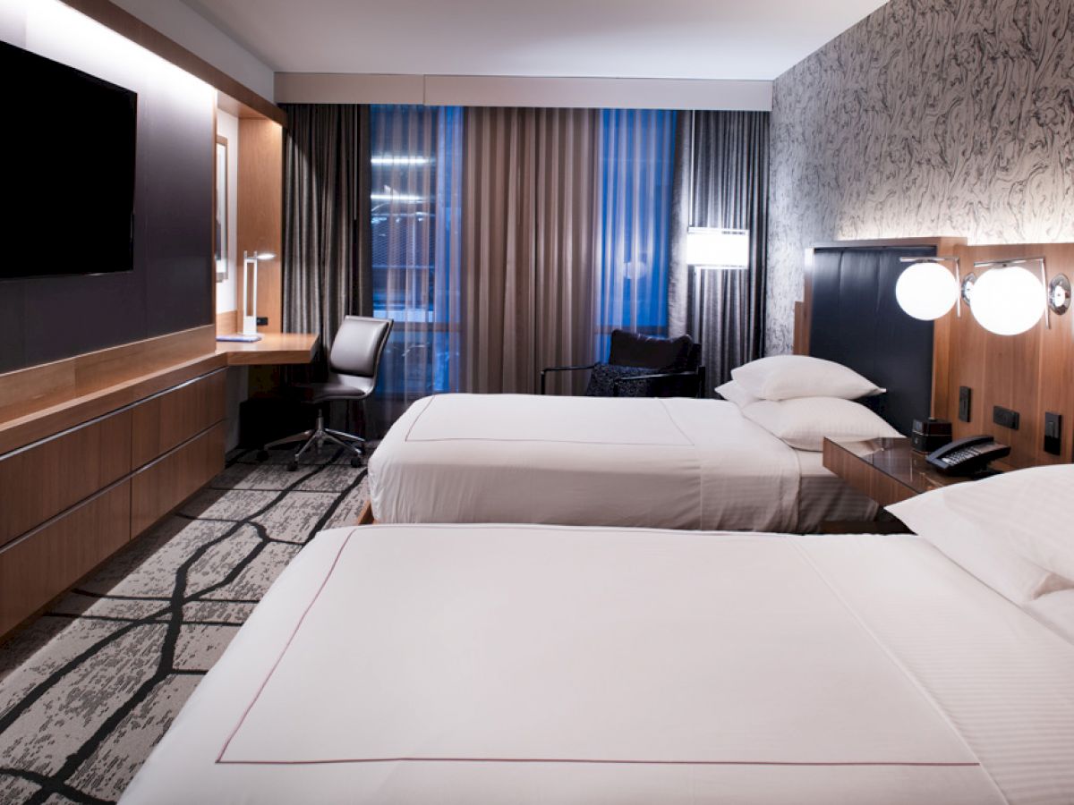 A modern hotel room with two double beds, a wall-mounted TV, a desk, and floor-to-ceiling windows with drapes, all lit by contemporary lighting fixtures.
