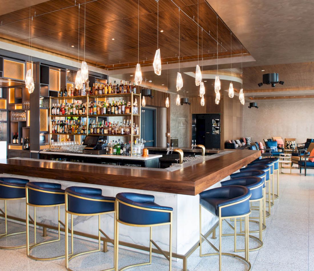 A wide view of a bar with a long counter, blue stools, and shelves of liquor behind it.