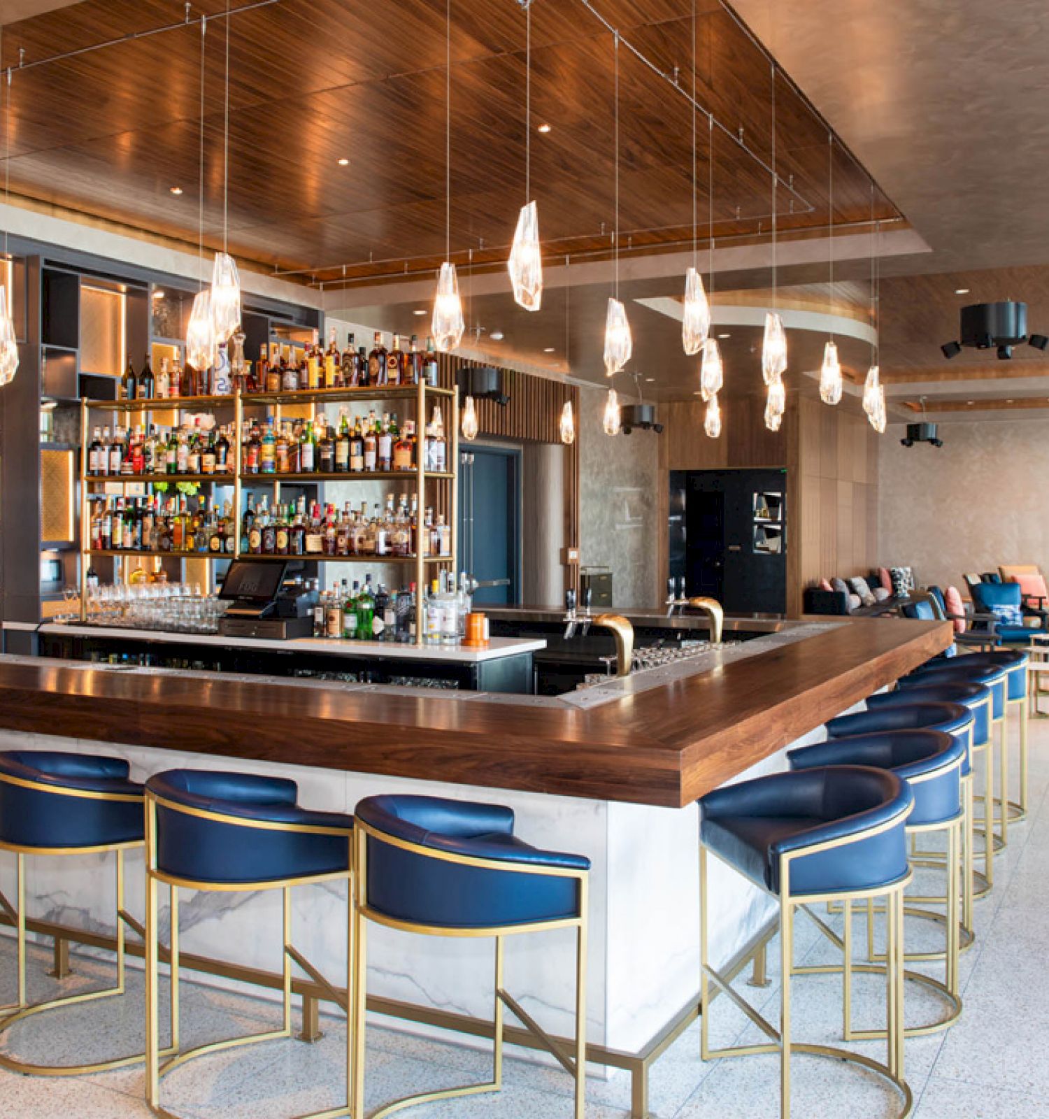 The image shows a modern bar with a sleek wooden counter, high stools, hanging lights, and a well-stocked array of alcohol bottles behind the bar.
