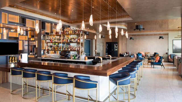 A modern bar with a long wooden counter, blue stools, and gold accents.
