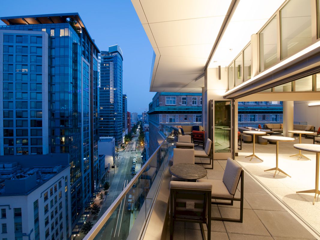 A modern rooftop terrace with seating, overlooking a city skyline with illuminated high-rise buildings at dusk.