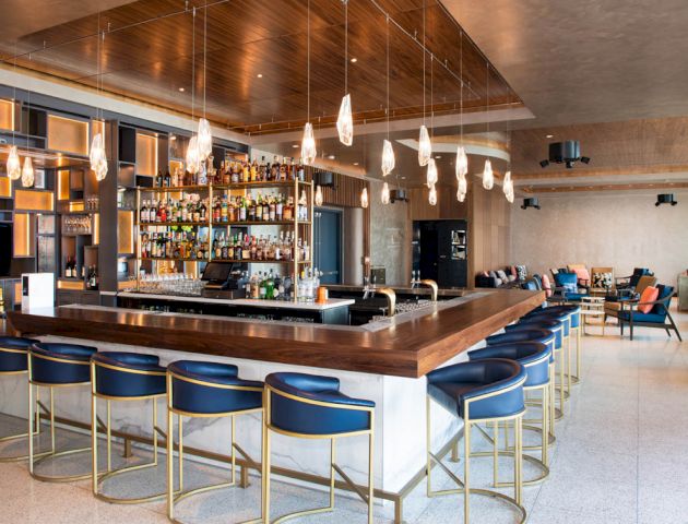 The image shows a modern bar with stylish seating, a well-stocked back shelf, pendant lights, and a cozy seating area in the background.