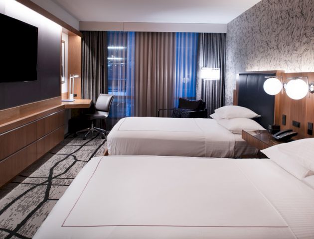 This image shows a modern hotel room with two beds, a wall-mounted TV, a desk with a chair, a patterned rug, and stylish decor.