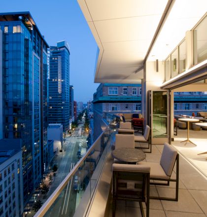 This image shows a modern cityscape viewed from a high-rise building's balcony or terrace at dusk with outdoor seating and table arrangements.