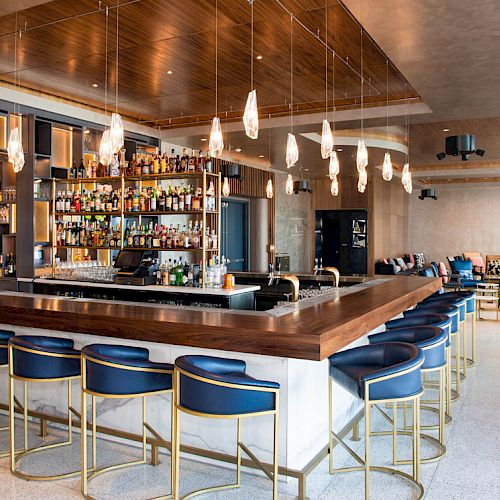 A modern bar with blue stools, a wooden counter, hanging lights, and a fully stocked shelf of bottles. The background shows a seating area.