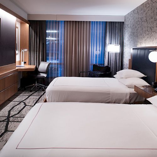 A modern hotel room with two beds, a desk with a chair, a wall-mounted TV, and stylish decor featuring patterned wallpaper and a large window.