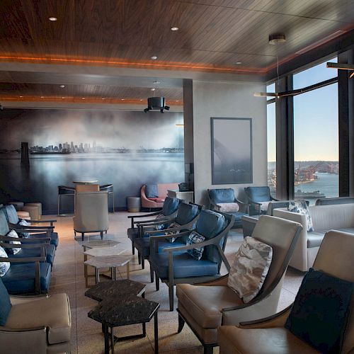 The image shows a modern lounge area with comfortable seating, a cityscape mural, large windows with a view of water, and a Ferris wheel outside.