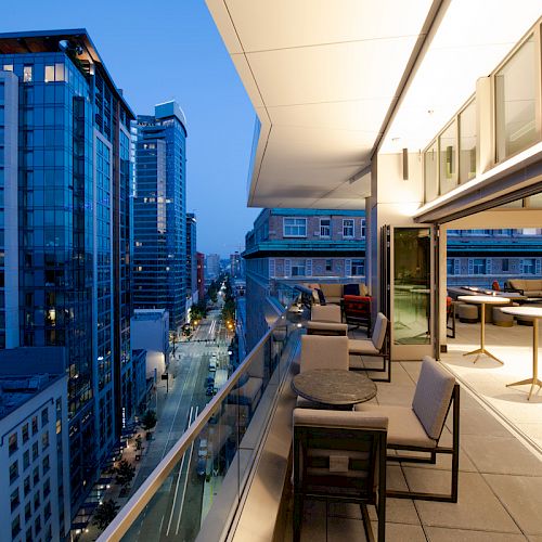 A modern cityscape at dusk with high-rise buildings and a rooftop terrace, featuring outdoor seating and tables, overlooking the city street below.