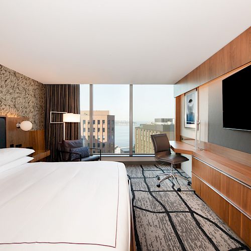 A modern hotel room with a large bed, wall-mounted TV, and city view through a wide window; a desk and chair are placed by the window.