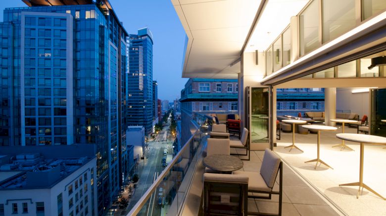 Modern high-rise buildings with a furnished balcony offering a city view at dusk; glass railings and an open indoor lounge area.