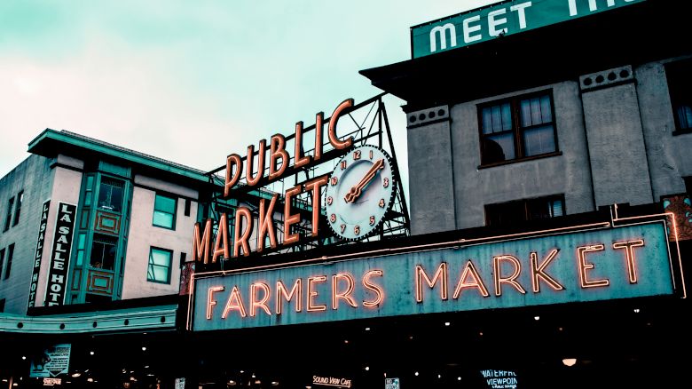 The image shows the iconic signage of Pike Place Market, a famous public market in Seattle, with "Public Market" and "Farmers Market" in neon lights.