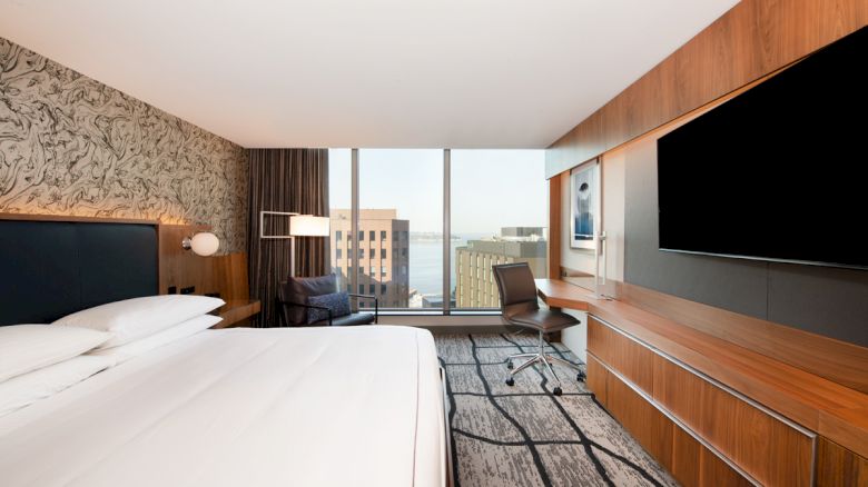 This image shows a modern hotel room with large windows, a bed, a desk with a chair, a TV, and a view of buildings outside the window.