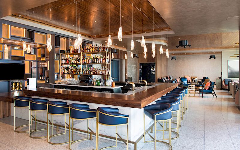 The image shows a modern bar with blue and gold bar stools, a variety of liquor bottles on shelves, hanging pendant lights, and a spacious, stylish seating area.