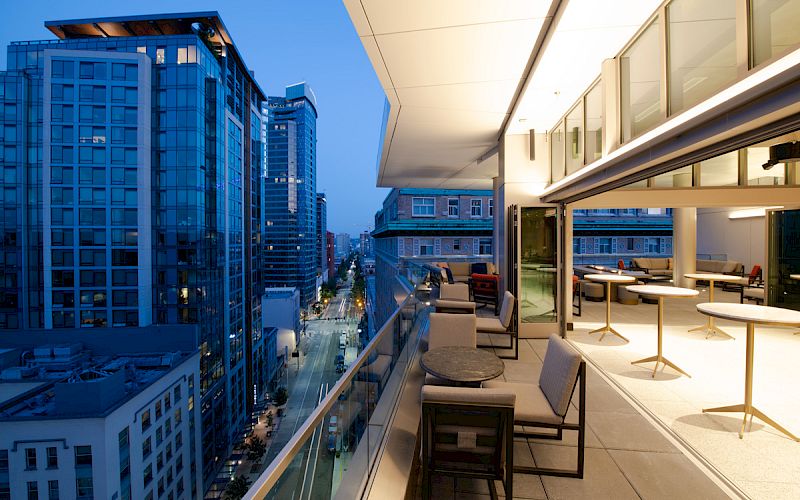 This image shows a modern cityscape at dusk featuring a rooftop patio with tables and chairs. High-rise buildings line the street below.