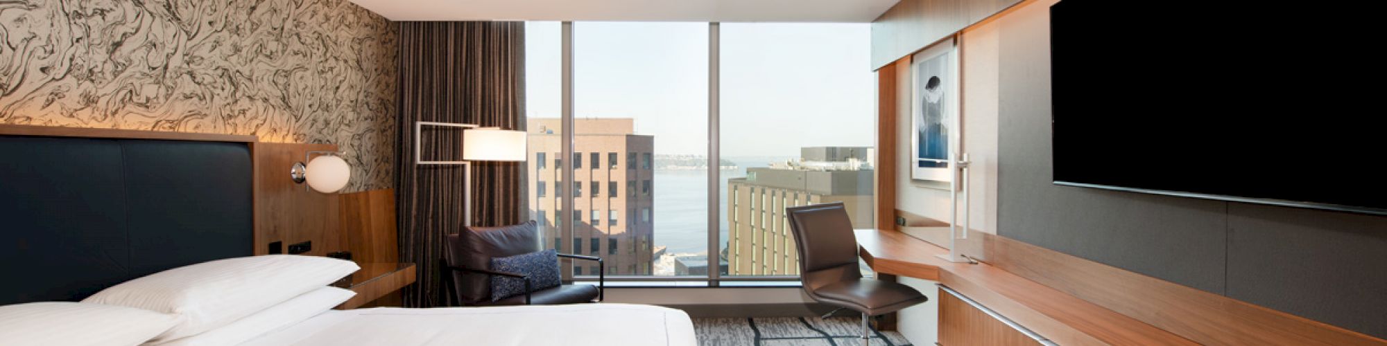 A modern hotel room with a large bed, a wall-mounted TV, a desk with a chair, and a floor-to-ceiling window with a cityscape view.
