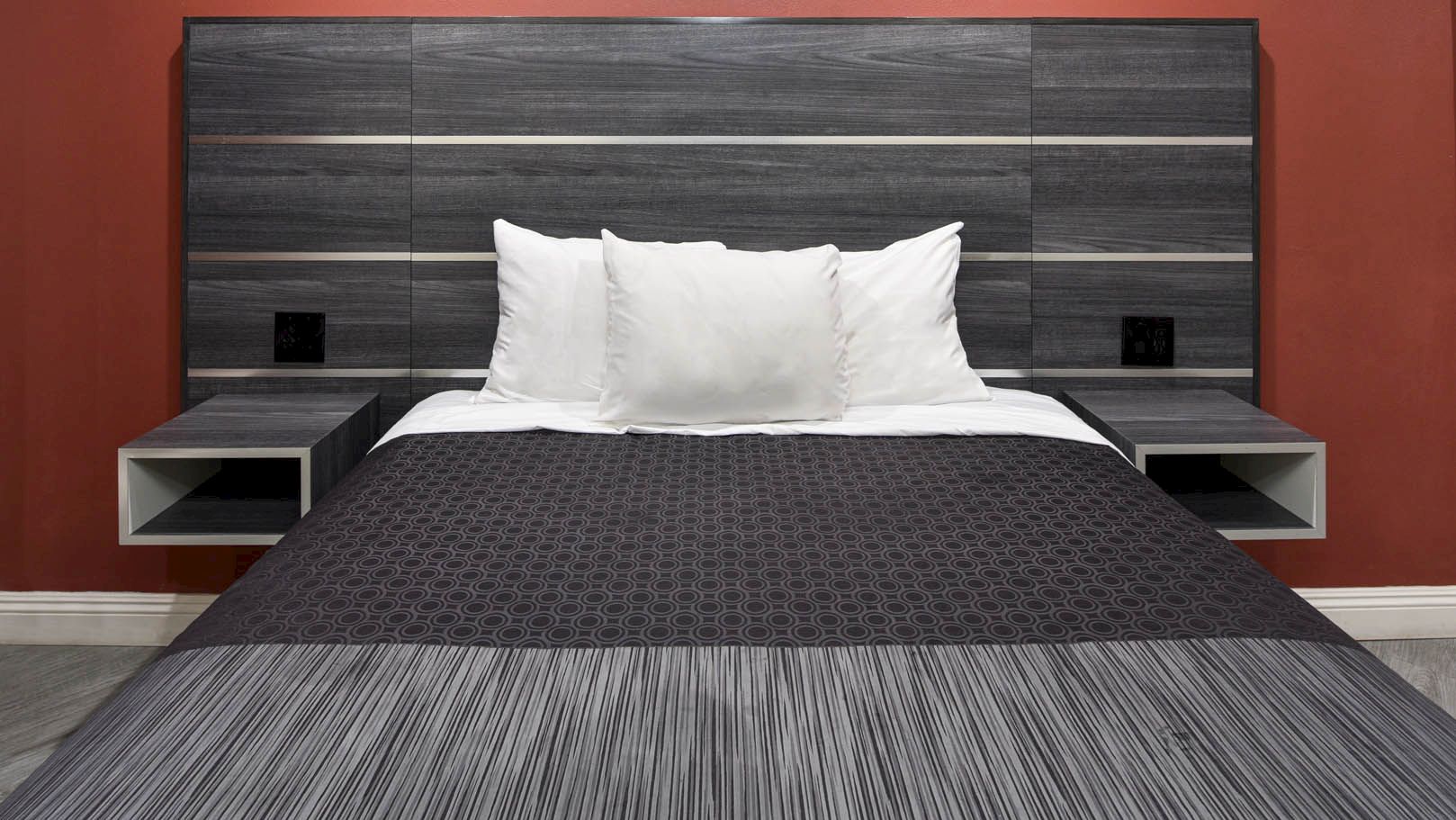 A neatly made bed with white pillows, dark bedding, and a modern headboard against a red wall, flanked by floating nightstands.