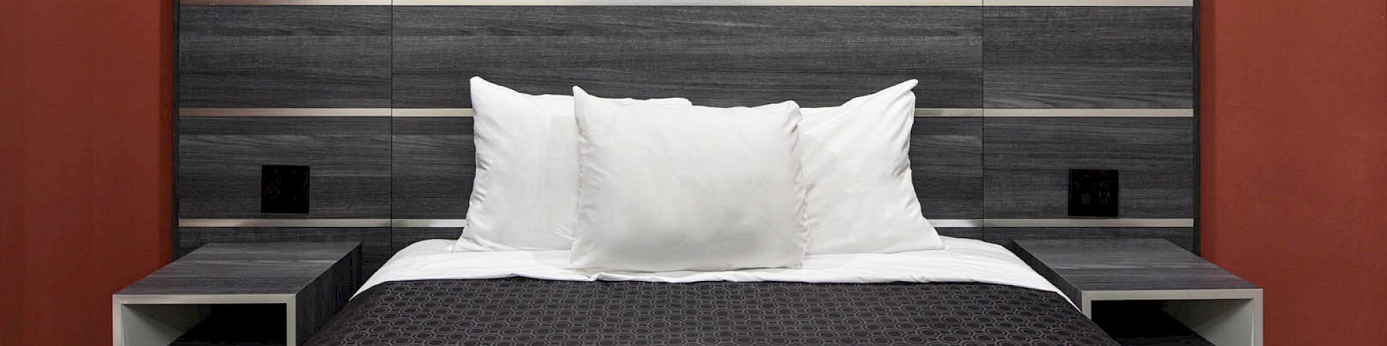 A neatly made bed with white pillows, dark bedding, and a modern headboard against a red wall, flanked by floating nightstands.